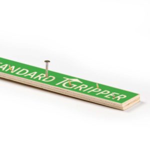 A metal nail half-hammered into a green and white yardstick labeled 'Standard Gripper' on a white background.