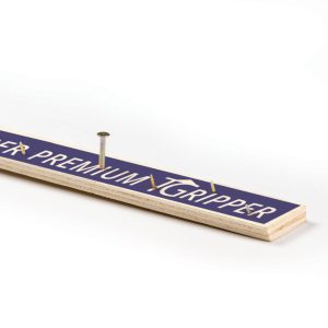 A wooden yardstick with 'GRIPPER PREMIUM' text and a nail partially driven through it, against a white background.